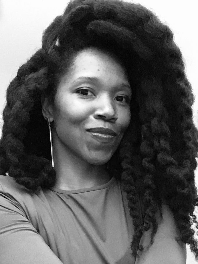 Black and white headshot of a woman with large, dark braided hair and dangling earrings, smiling with arms crossed.