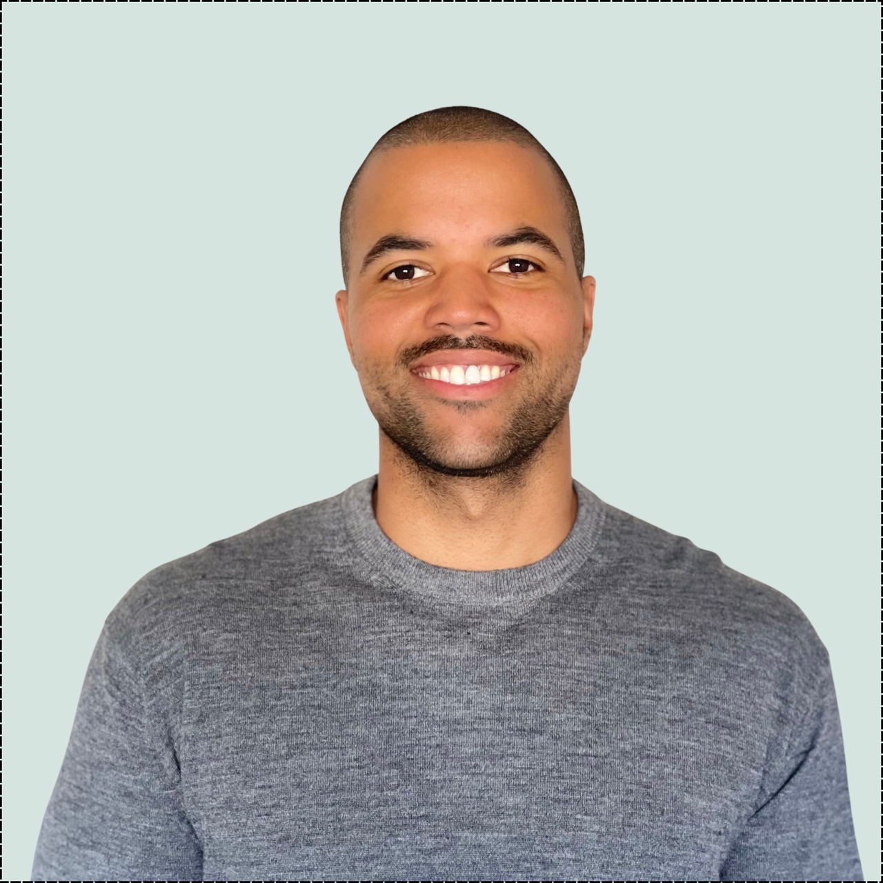 Smiling Black man with shaved head in gray sweater, against a light green background.
