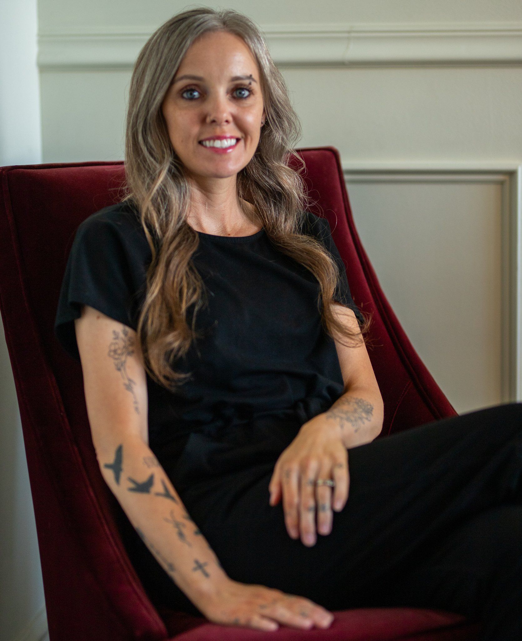 Woman with graying hair and tattoos smiles while sitting in a red chair.