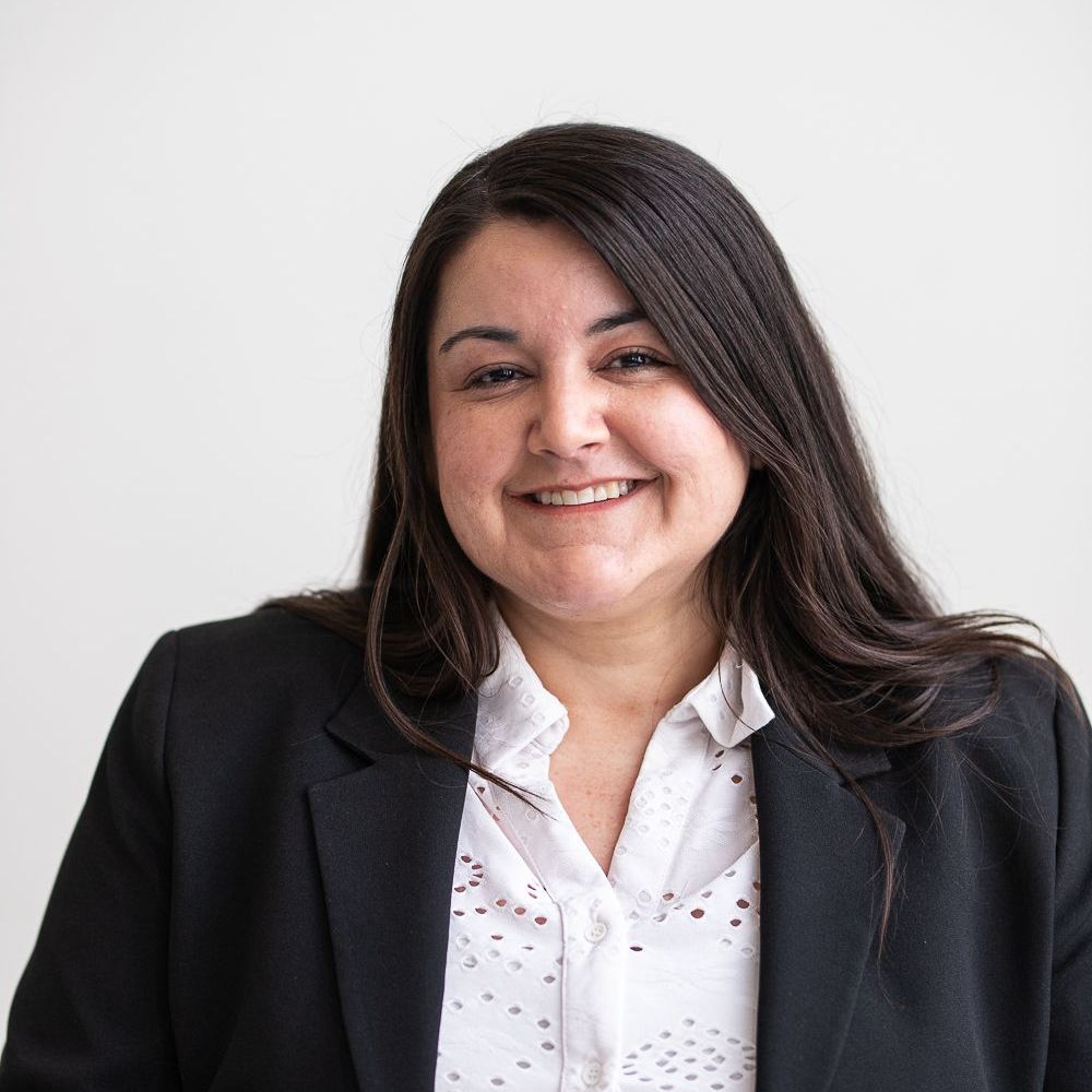 Woman with long dark hair, wearing a white blouse and black blazer, smiling at the camera.
