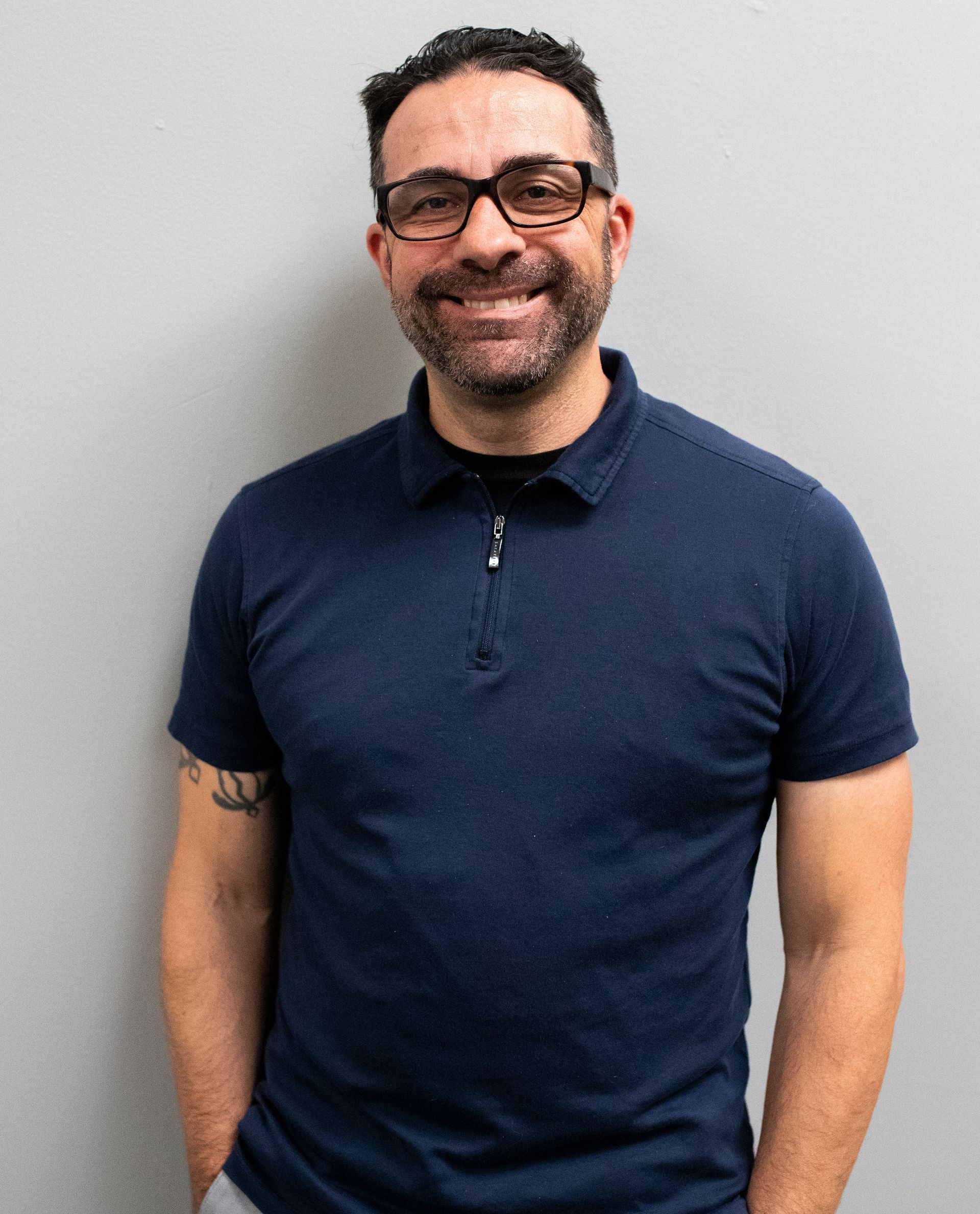Man with glasses, dark hair, and a beard, wearing a blue polo shirt and smiling, standing in front of a gray wall.