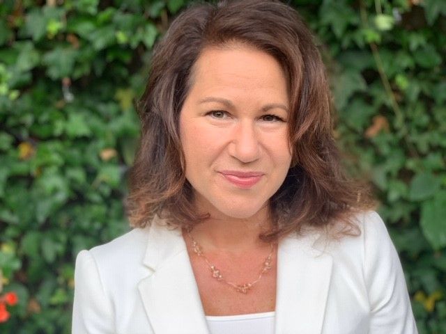 Woman with brown hair wearing a white blazer and necklace smiles in front of a green leafy background.