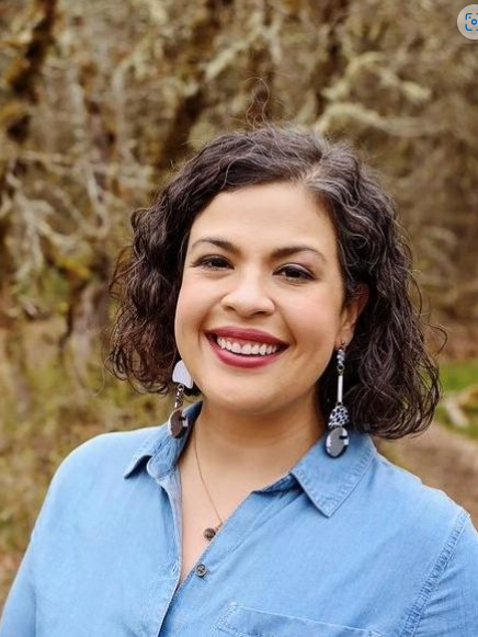 Woman with dark curly hair smiles at the camera, wearing a blue shirt and dangling earrings, with a blurred outdoor background.