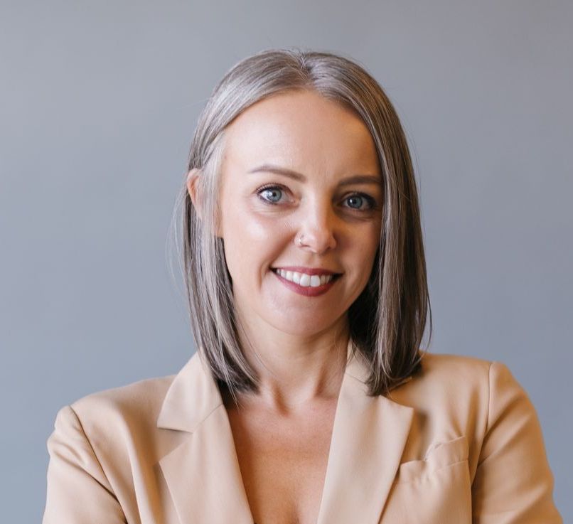Woman with graying hair, wearing a tan blazer, smiling at the camera, set against a neutral gray backdrop.