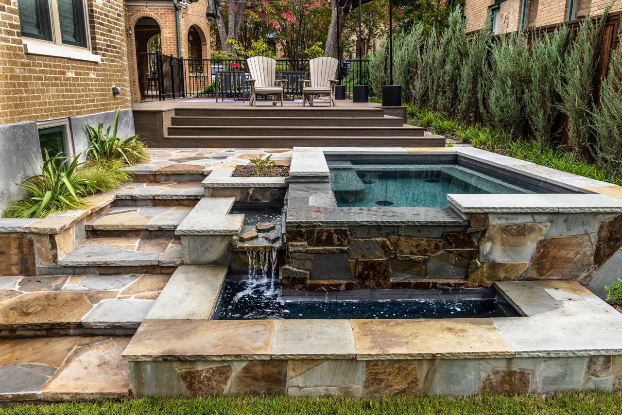 Stone patio with a tiered design featuring a spa and waterfall, leading to a raised deck with seating.