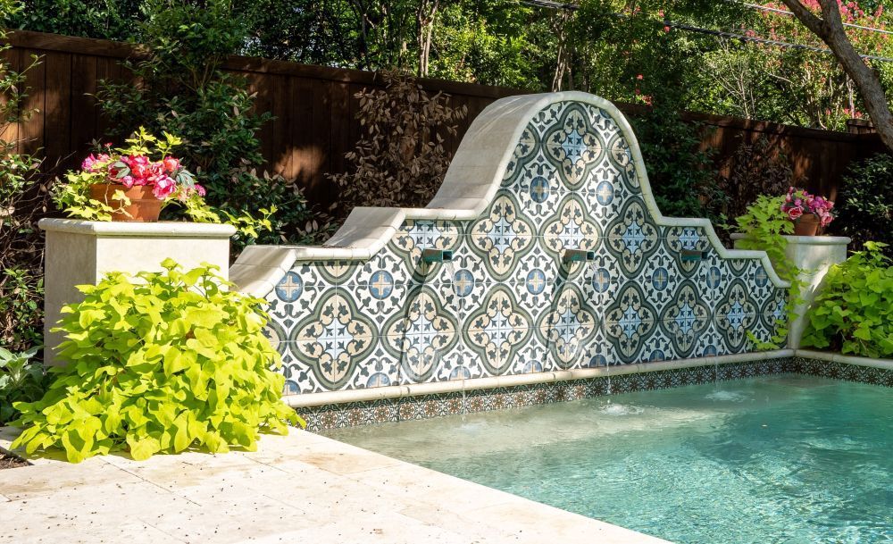 Pool with a tiled fountain against a wooden fence, with plants on each side.