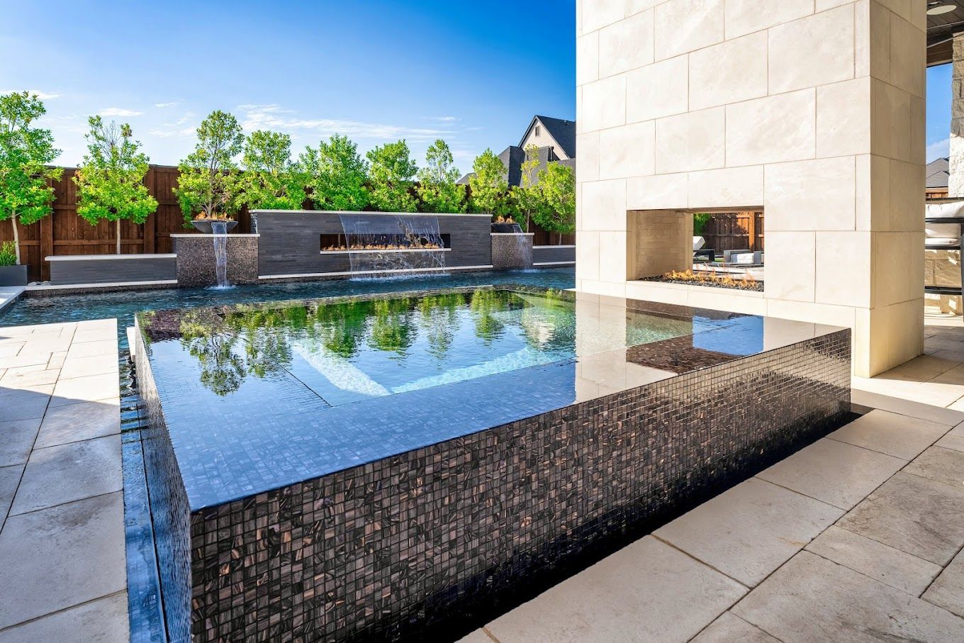 Luxurious outdoor pool with tiled walls, fireplace, and green trees.