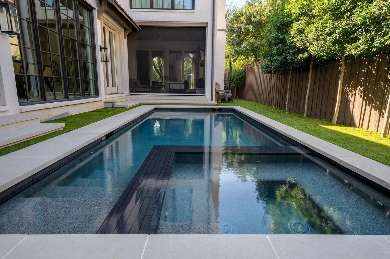 Rectangular pool with dark trim, stairs, and patio, alongside a house and grass.
