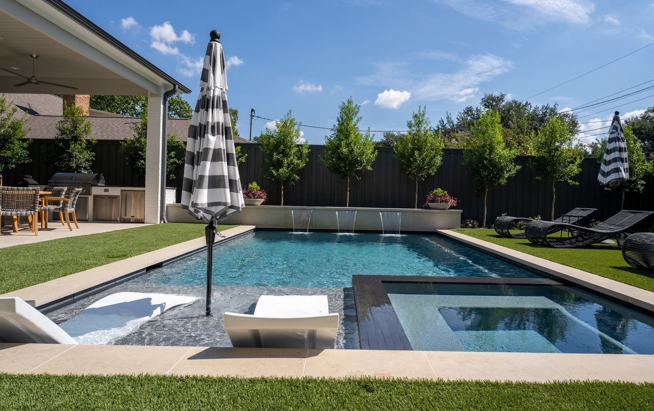 Backyard pool with umbrellas, lounge chairs, and a small waterfall feature on a sunny day.