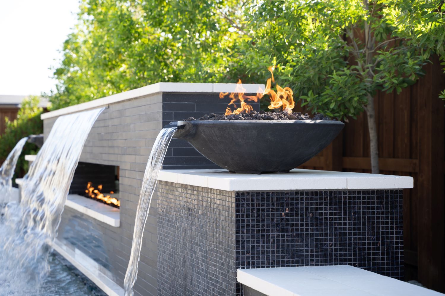 Water feature with waterfalls and a fire bowl, set in front of green foliage and a wooden fence.