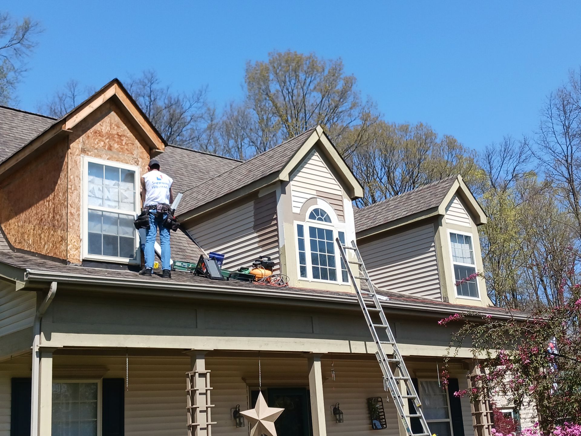 Man on roof repairing dormer siding on a two-story house. Ladder, tools, and blue sky visible.