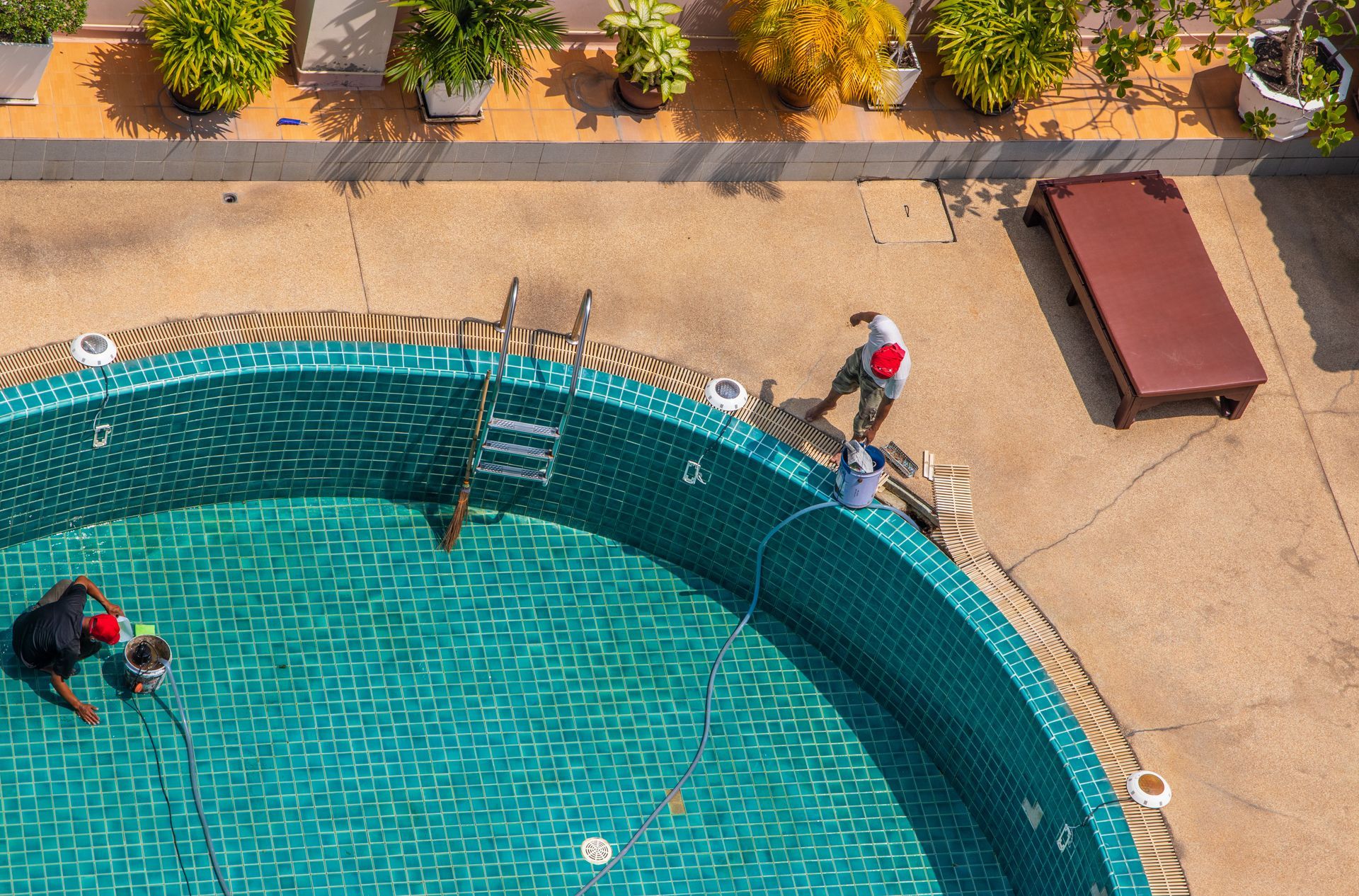 Overhead view of workers inside an empty, teal-tiled swimming pool on a patio with plants and a lounge chair.