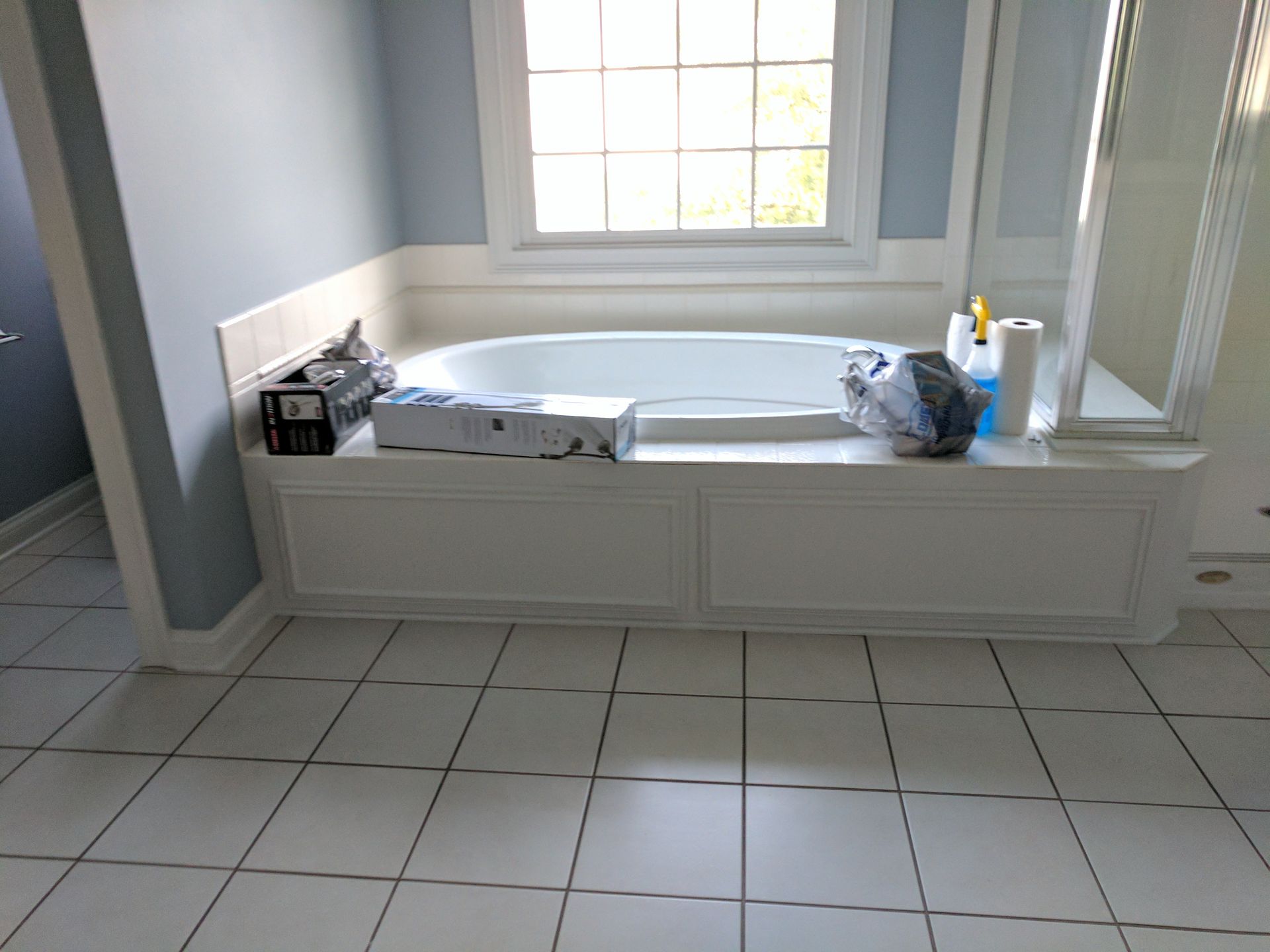 White-tiled bathroom with a built-in tub, window, and various items on the tub's ledge.