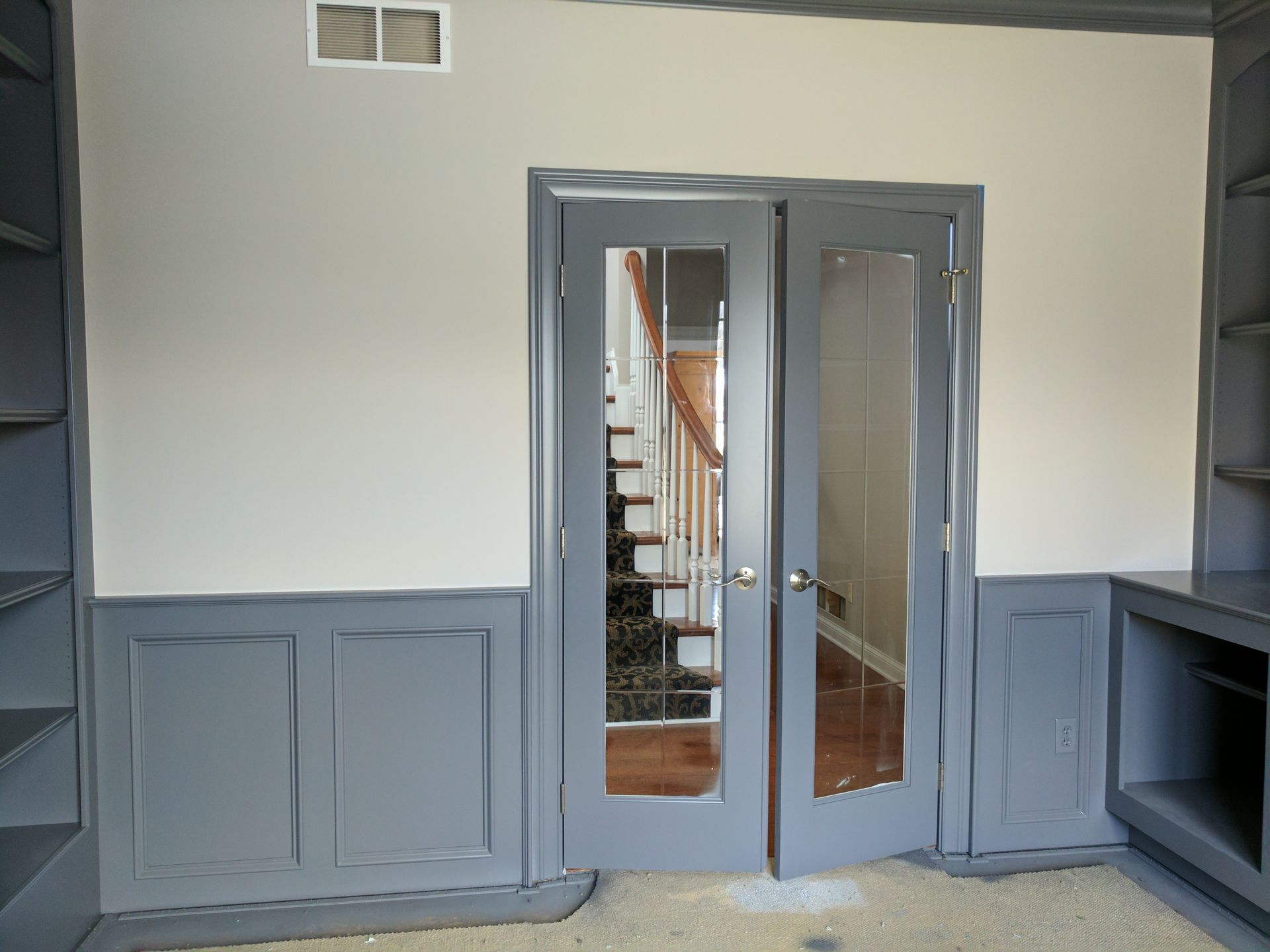Gray French doors, wainscoting, and bookshelves in a room with a staircase visible through the doors.