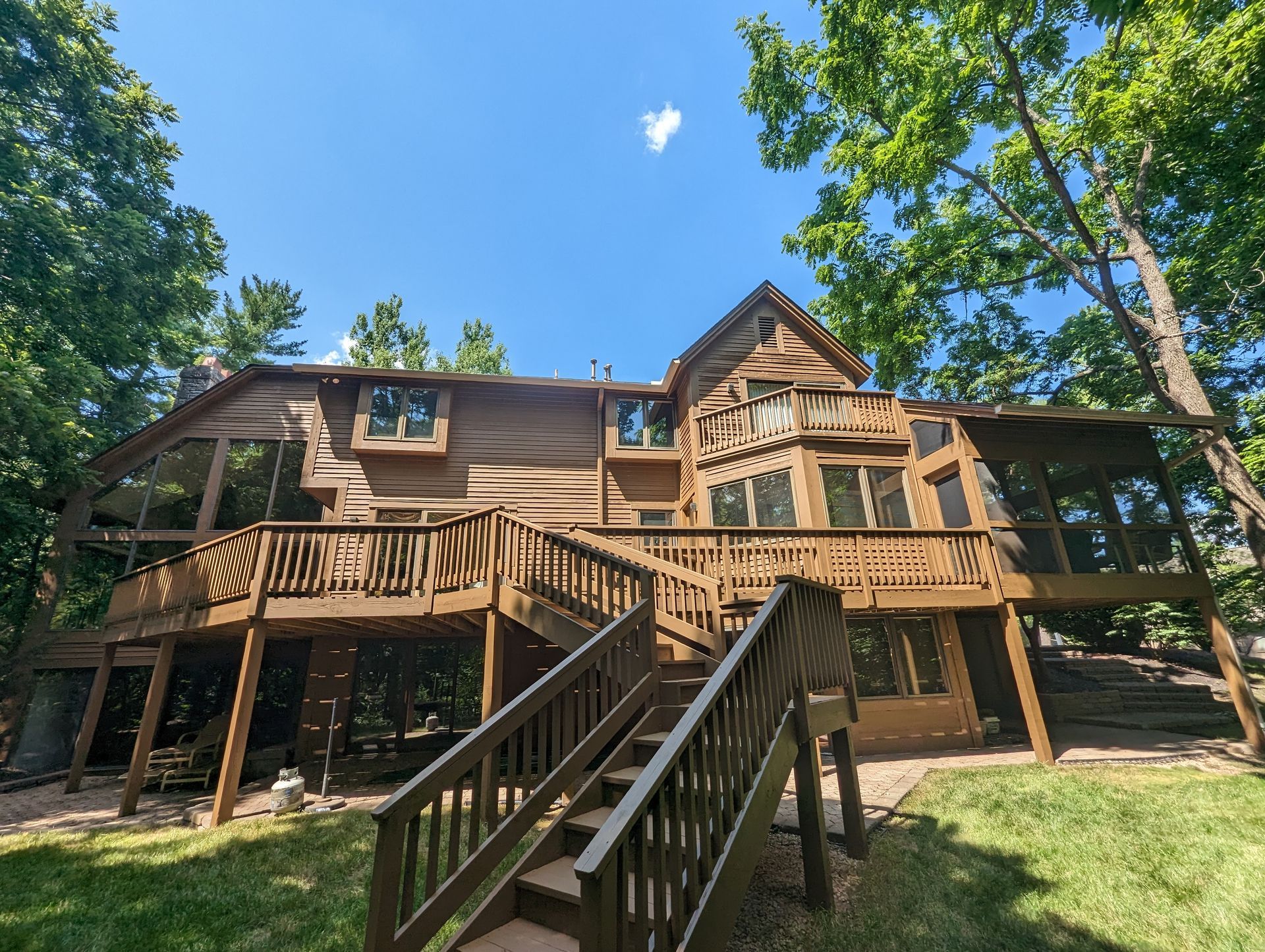 Wooden house with a large deck, surrounded by trees, on a sunny day.