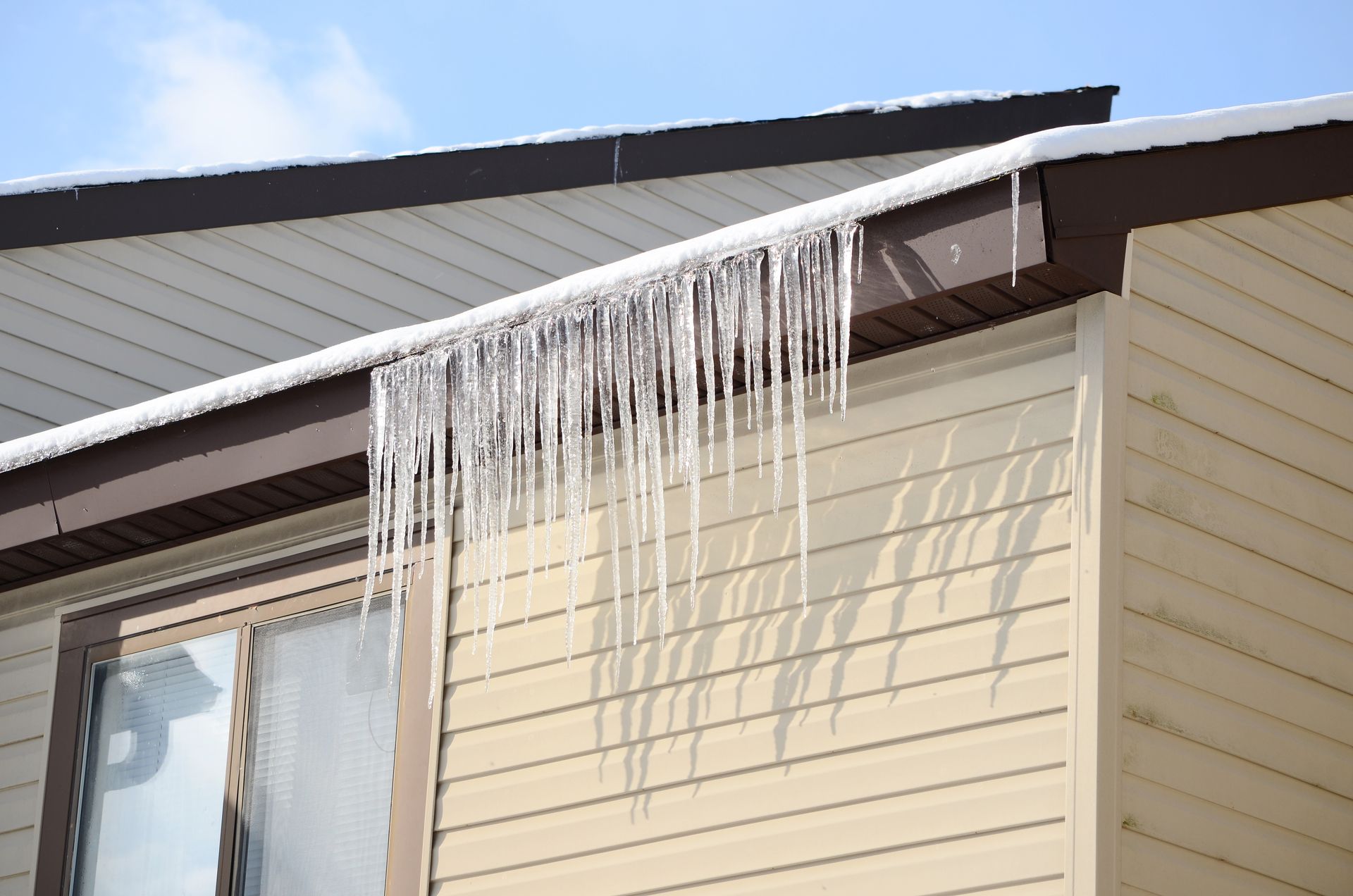 Icicles hang from a brown-trimmed roofline on a light tan building with a window.