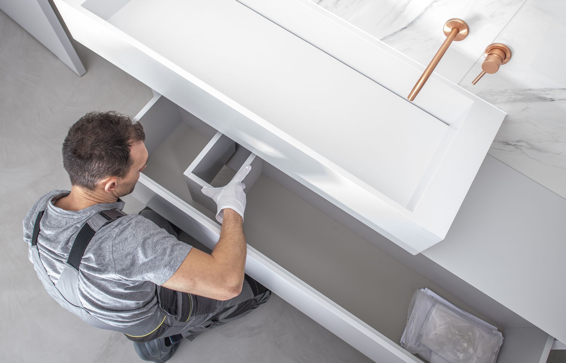 Man in work clothes installing a white bathroom vanity with a drawer, copper faucet nearby.