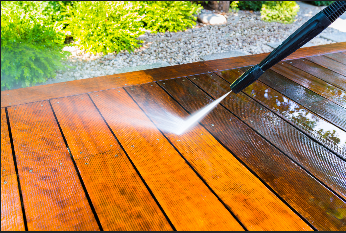 Pressure washer spraying water onto a wooden deck, cleaning the wood and revealing its natural color in an outdoor setting.