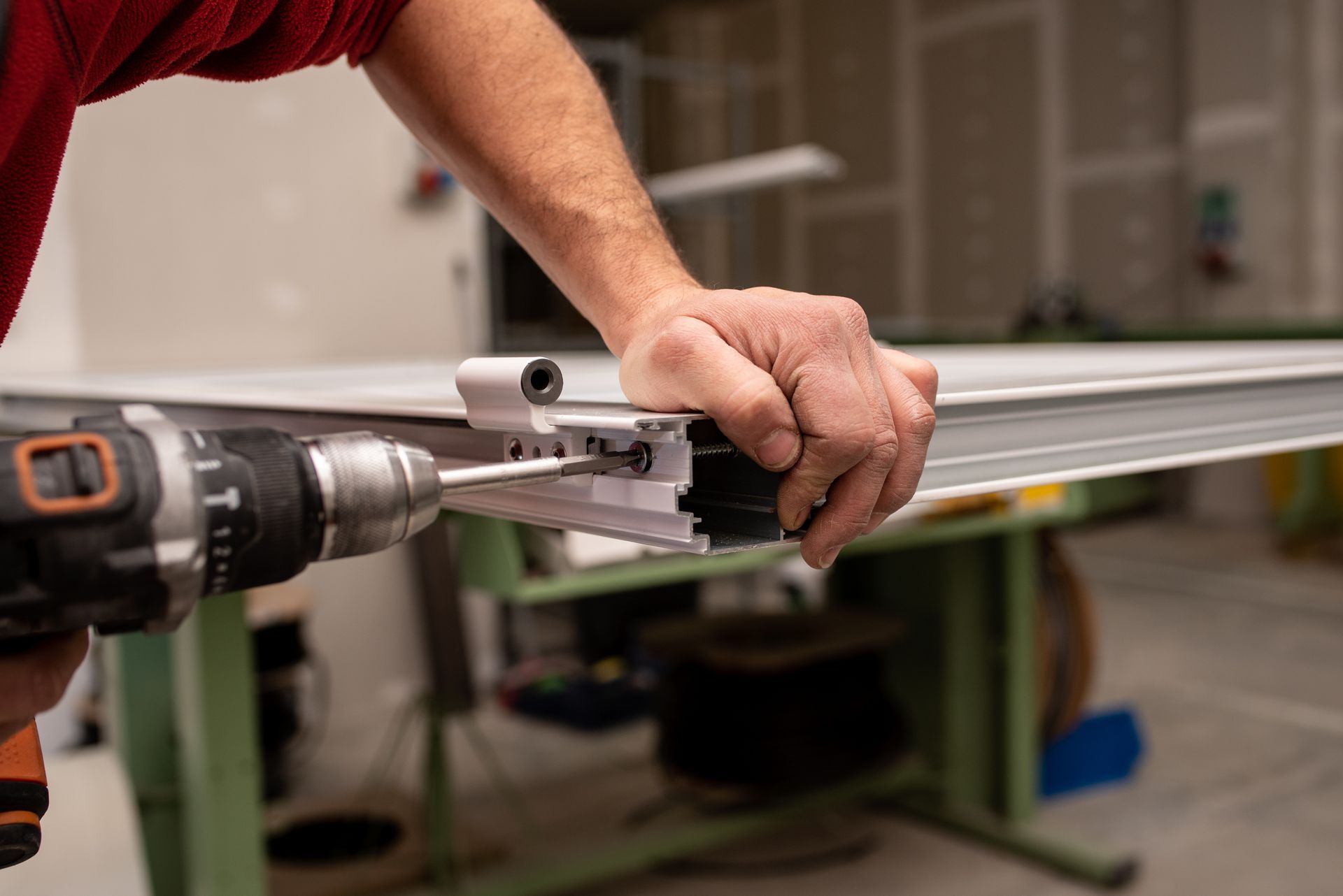 A person using a power drill to screw a metal frame together on a work table inside a workshop.
