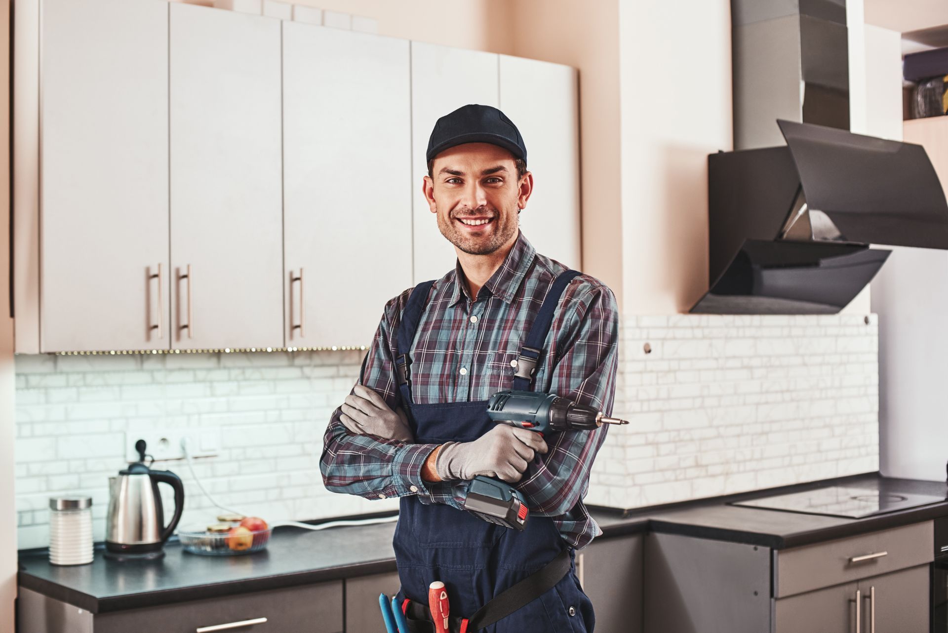 Man in work clothes holding a drill, standing in a kitchen, smiling.