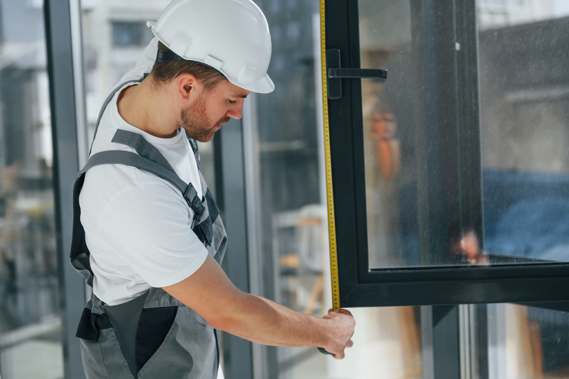 A construction worker in a white hard hat and overalls measures a window with a tape measure.