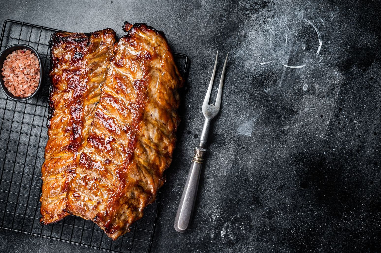 Glazed rack of ribs on a wire cooling rack next to a small bowl of spices and a metal carving fork on a dark surface.
