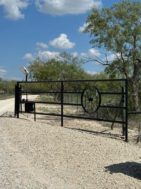 Black metal gate with circular emblem on a gravel drive, framed by trees under a blue sky with clouds