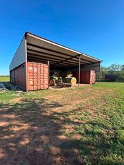 Large open-sided shed with farm equipment and shipping containers in a grassy field under a blue sky