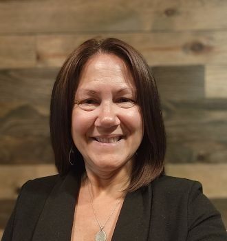 A woman in a black jacket and necklace is smiling in front of a wooden wall.