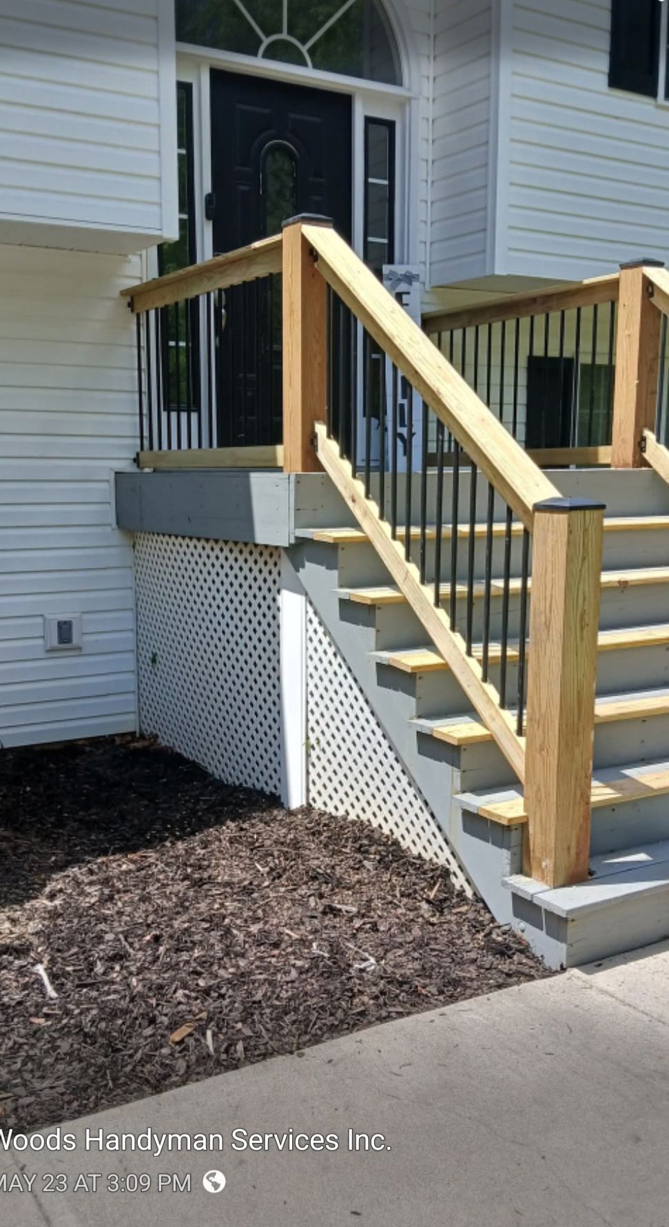 A newly built wooden front porch and stairs with black metal railings leading to a house with white siding.