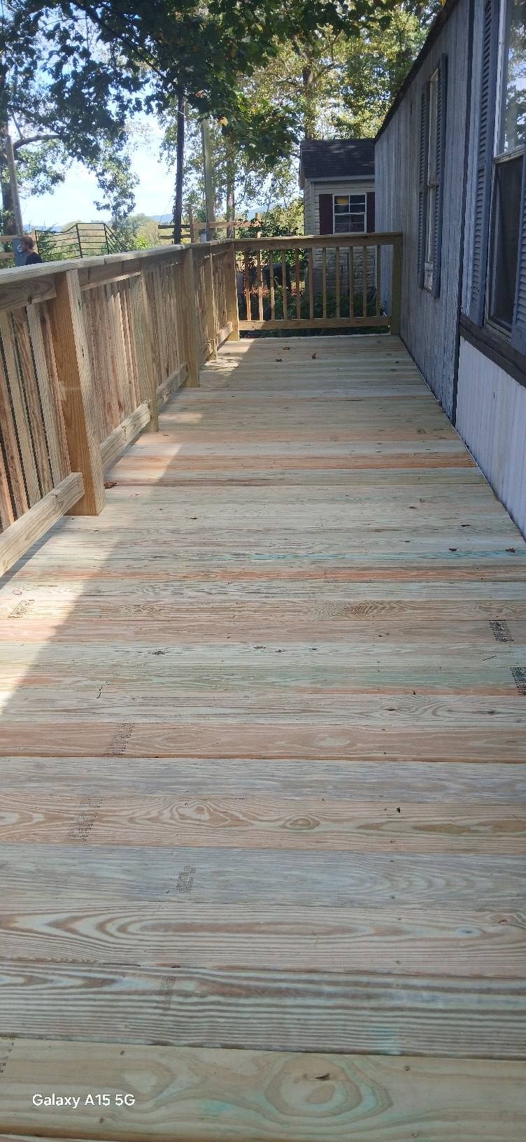 A close-up, low-angle view of a grooved wooden deck floor, with a wooden railing and trees in the background.