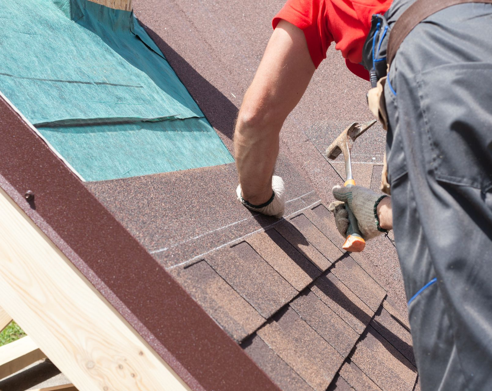 A worker in a hard hat and high-visibility vest uses tools while crouching to install shingles on a sloped roof.