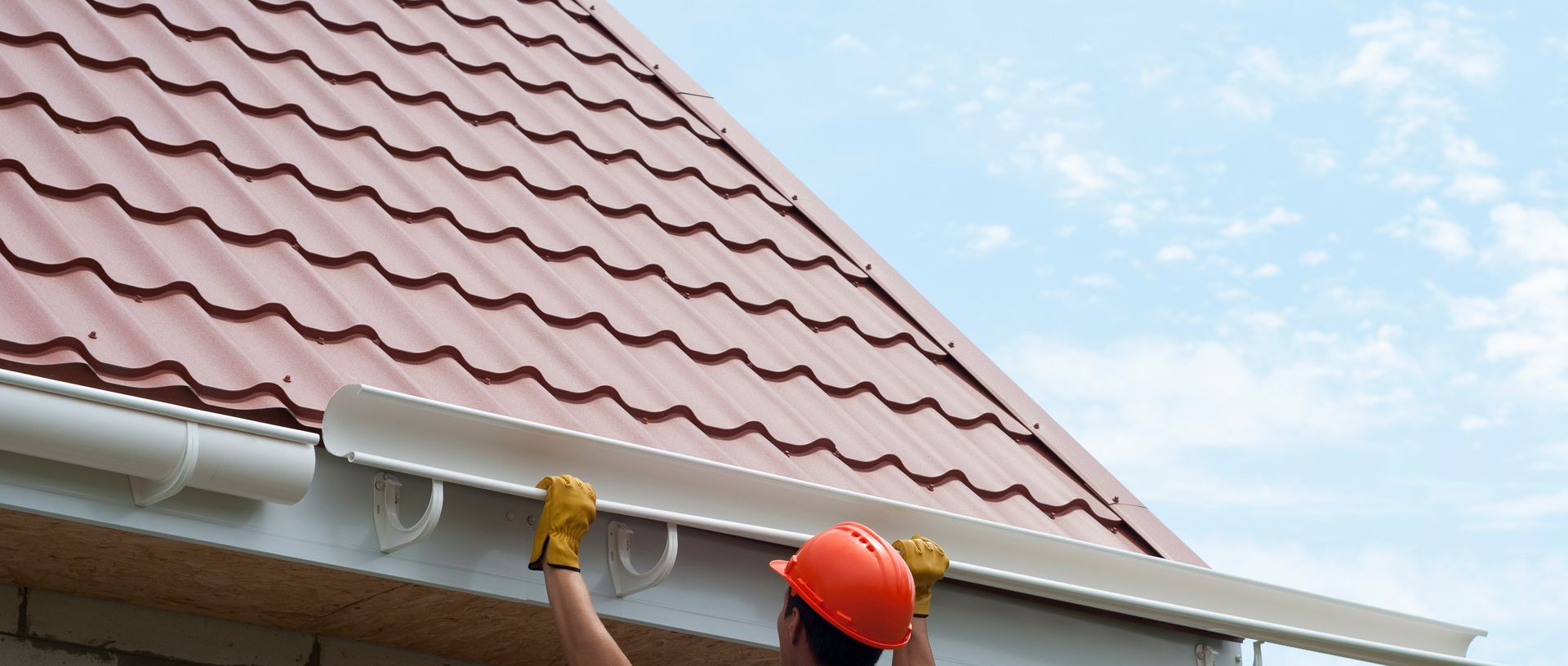 A professional in a blue uniform and gloves uses a ladder to inspect or clean the gutters of a metal roof.