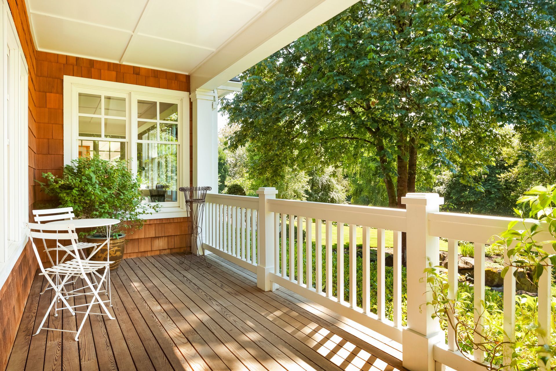 A wooden porch with a small white metal table and two chairs, featuring a white railing overlooking lush green trees.