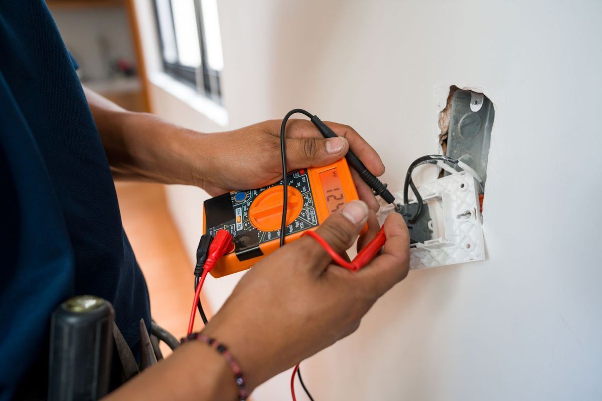 An electrician uses a digital multimeter to test the wiring of an exposed electrical outlet in a room.