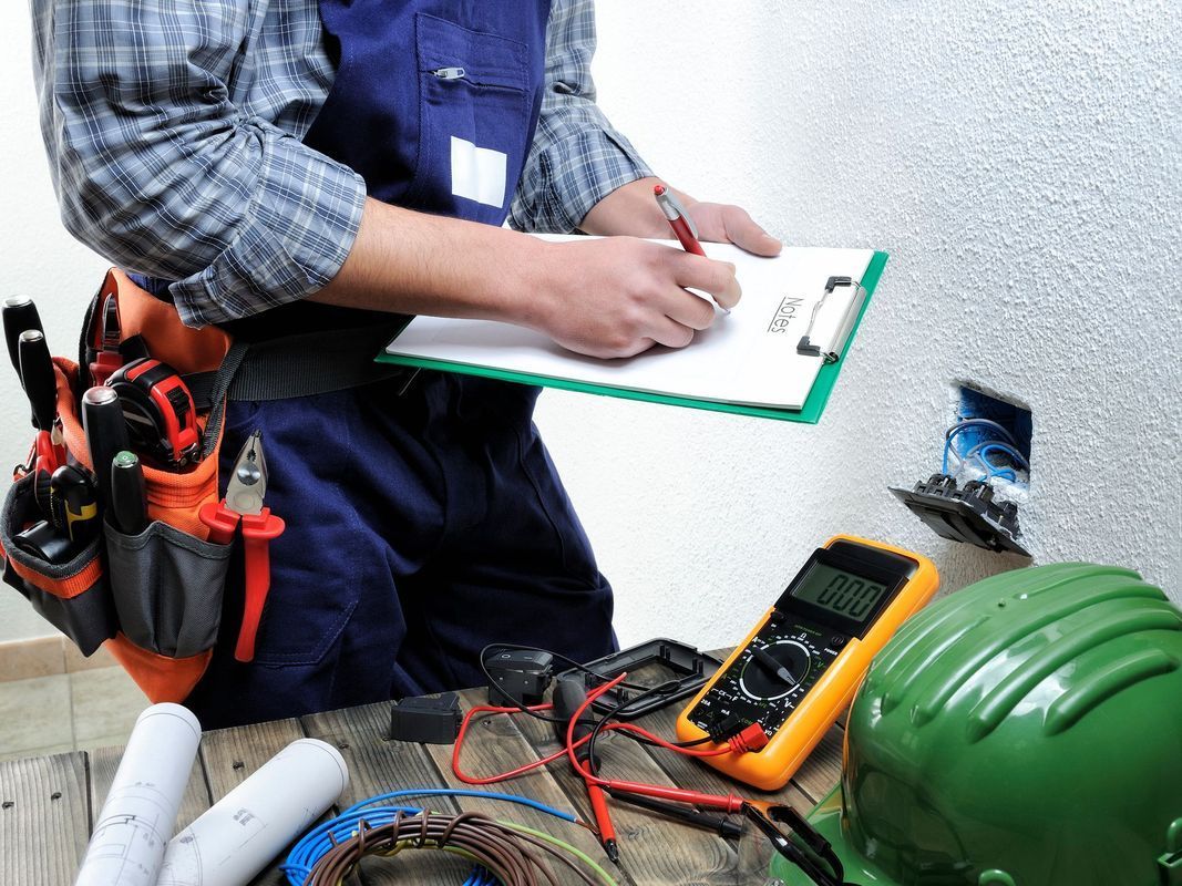 An electrician writes on a clipboard next to an open wall outlet, with tools, a multimeter, and a hard hat on a table.