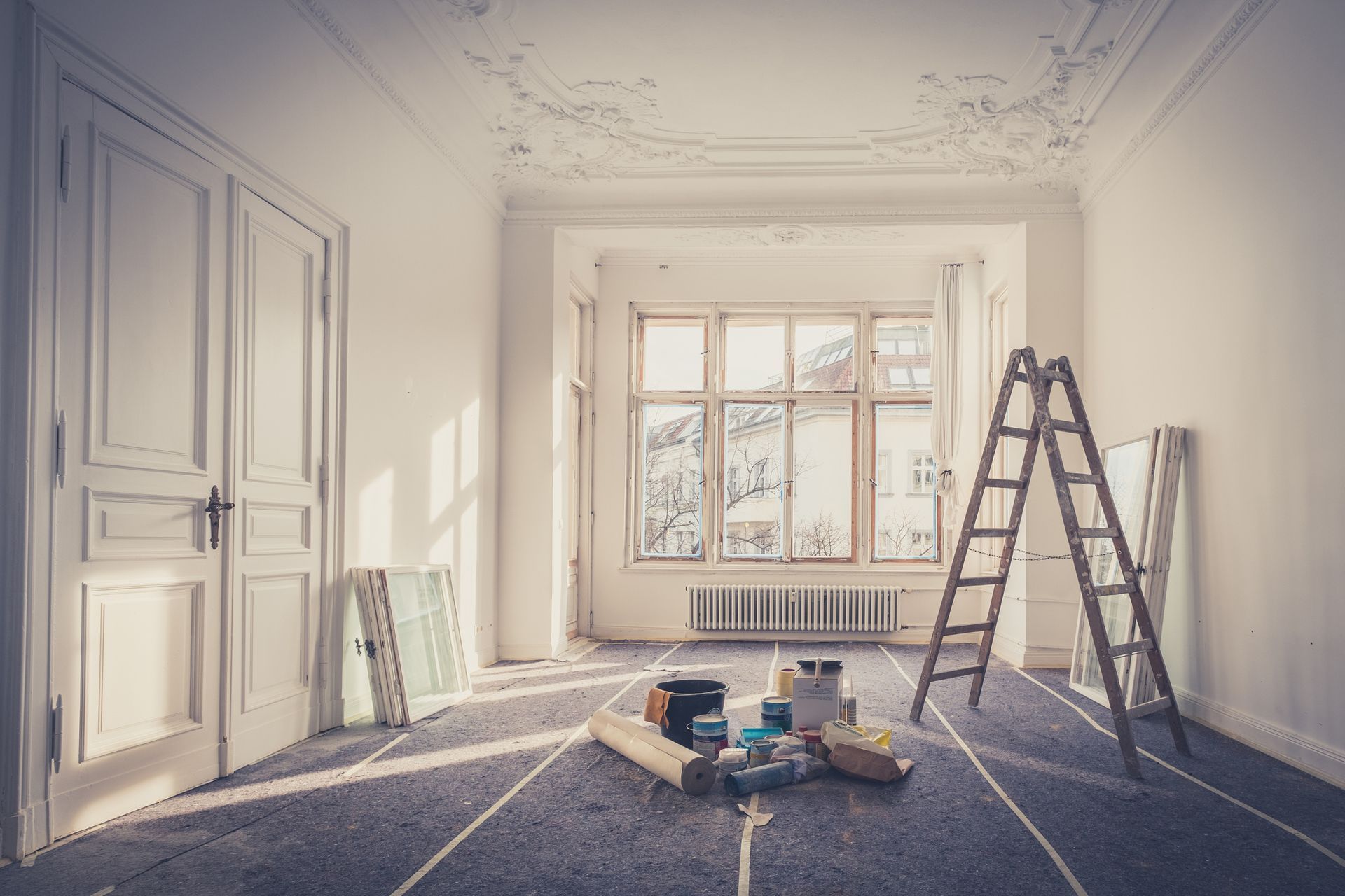 A bright room undergoing renovation with a wooden stepladder, painting supplies, and doors in a white, minimalist space.
