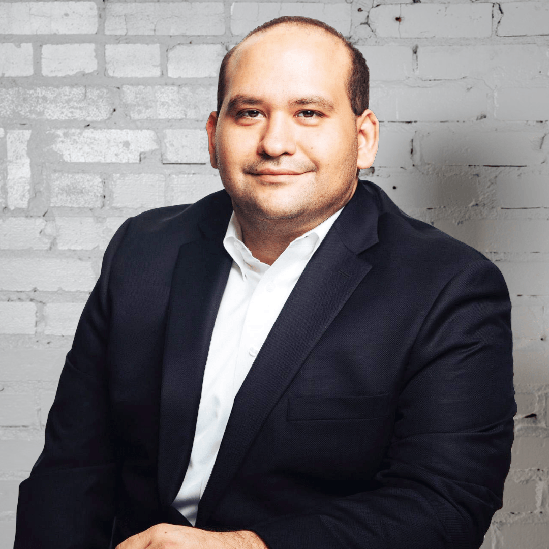 A man in a suit and white shirt is sitting in front of a brick wall.