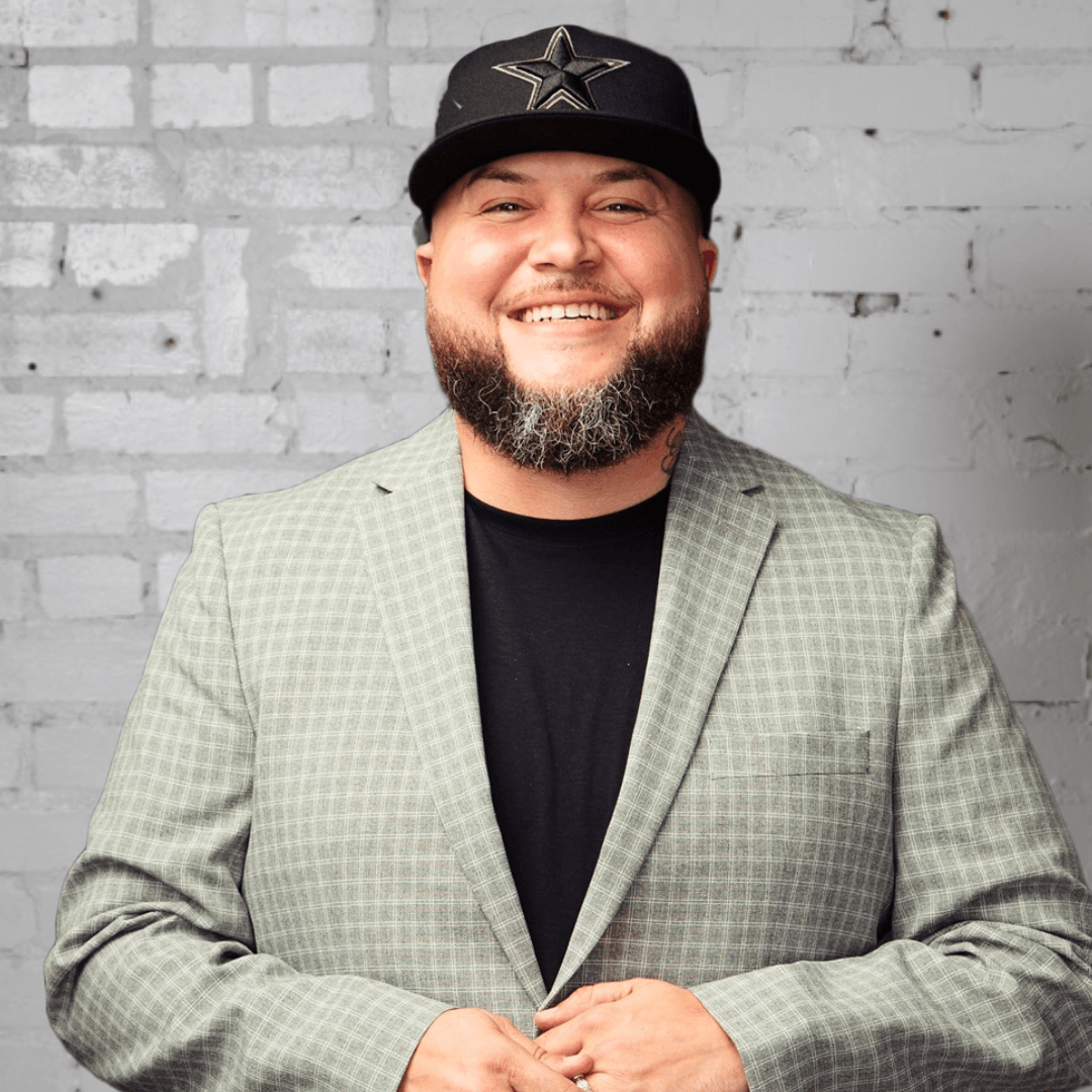 Juan Varela with a beard wearing a hat and a suit is smiling in front of a brick wall.