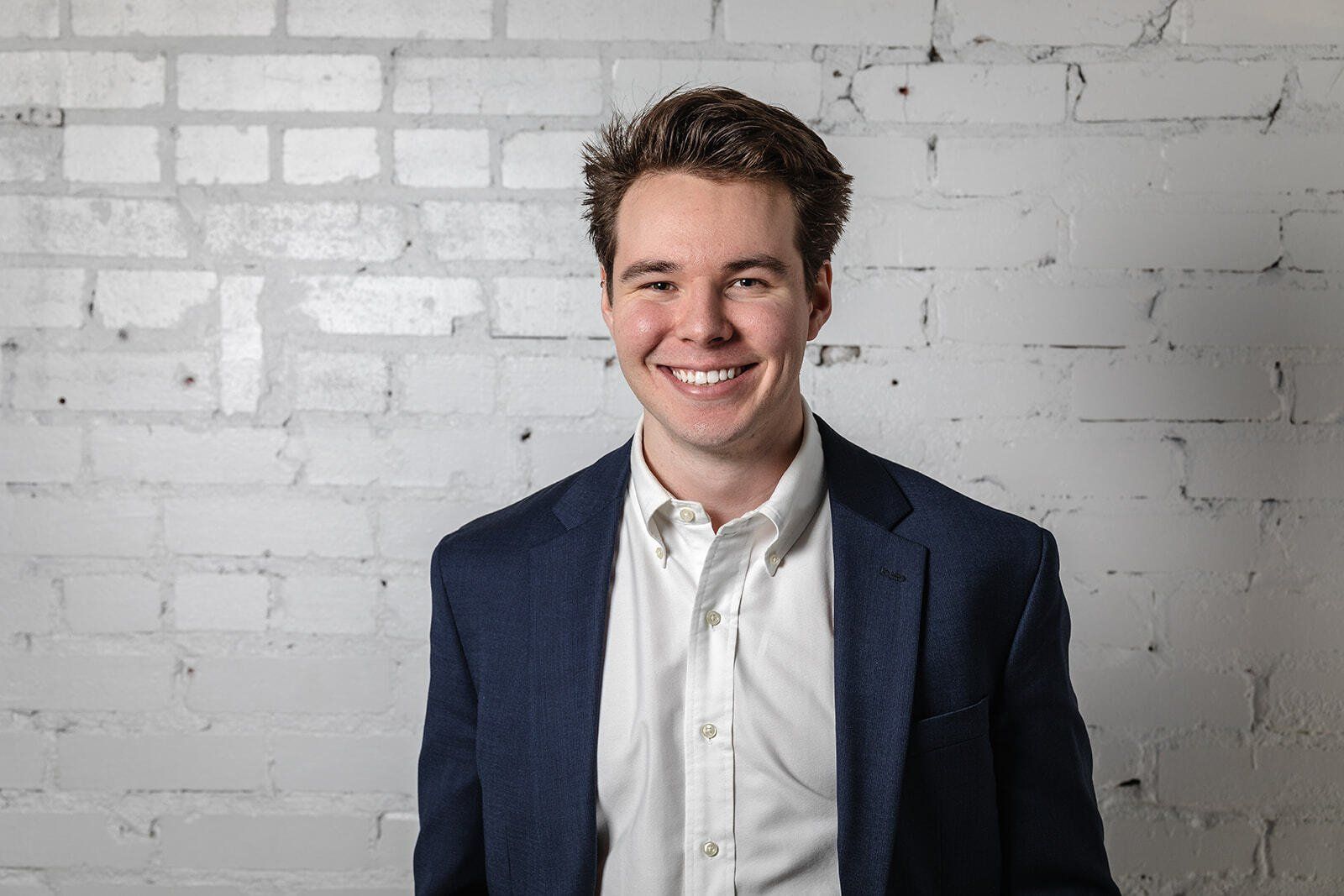 A man in a suit and white shirt is smiling in front of a white brick wall.