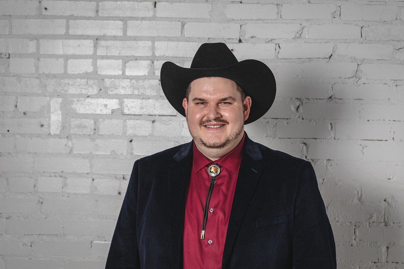 A man wearing a cowboy hat and a red shirt is standing in front of a white brick wall.