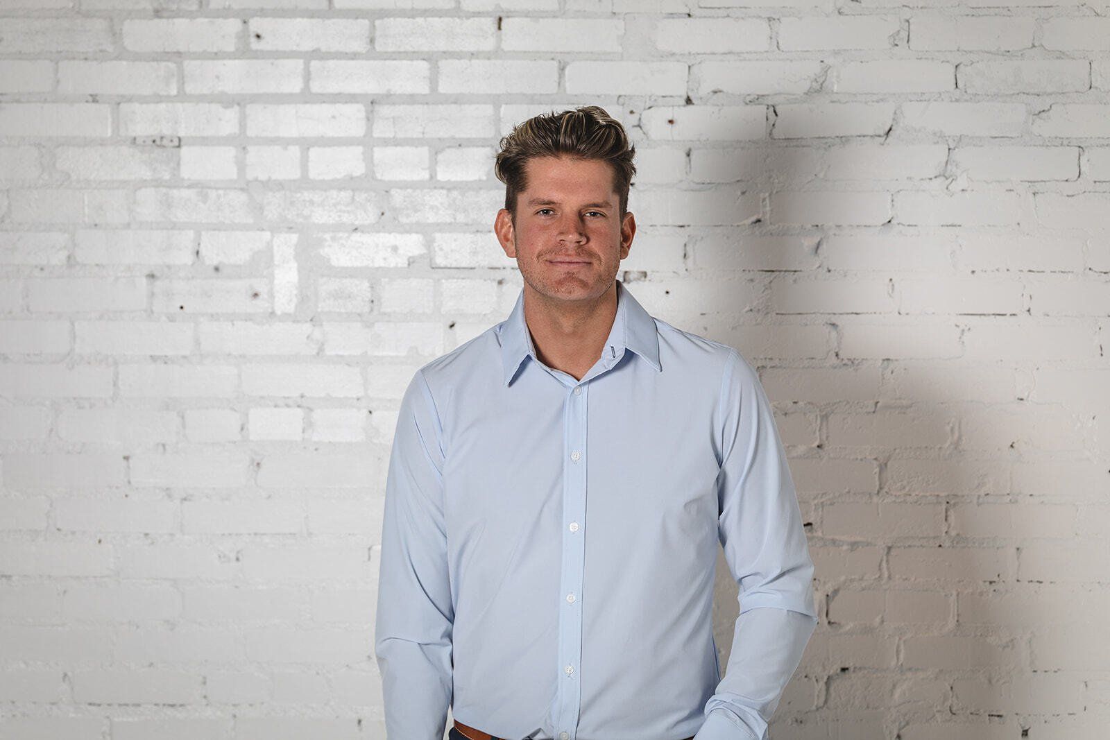 A man in a light blue shirt is standing in front of a white brick wall.