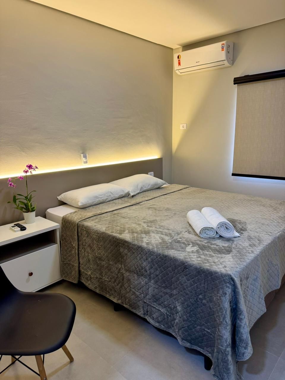 Um quarto de hotel arrumado com uma cama queen-size com colcha cinza estampada, duas toalhas brancas, uma mesa de cabeceira e iluminação de parede.