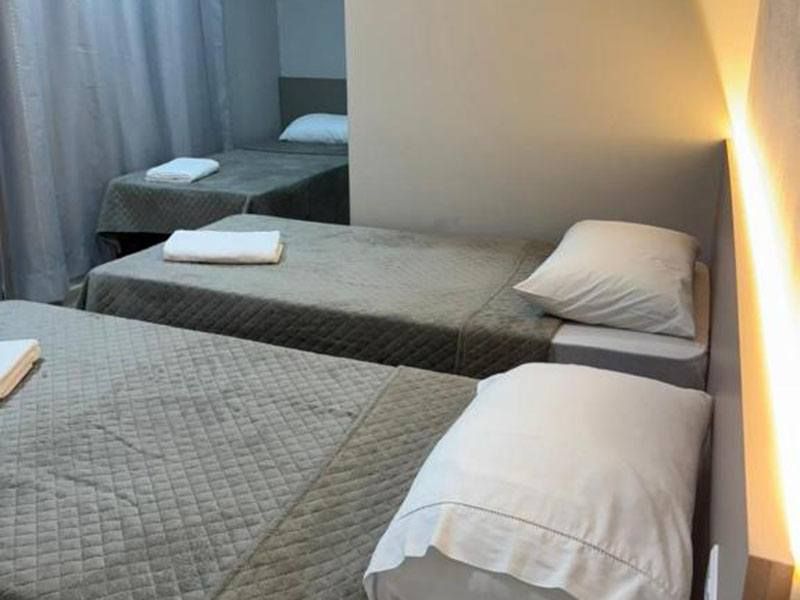 Um quarto com três camas de solteiro dispostas em fila, cada uma com uma colcha de cor clara, um travesseiro e uma toalha dobrada.