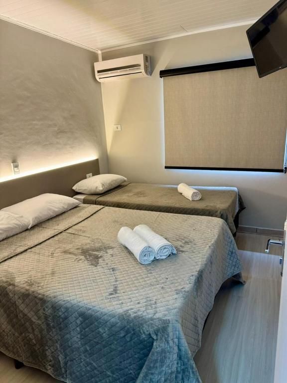 Um quarto de hotel limpo com uma cama de casal e uma cama de solteiro, ambas com lençóis brancos, ao lado de uma janela com persiana bege.