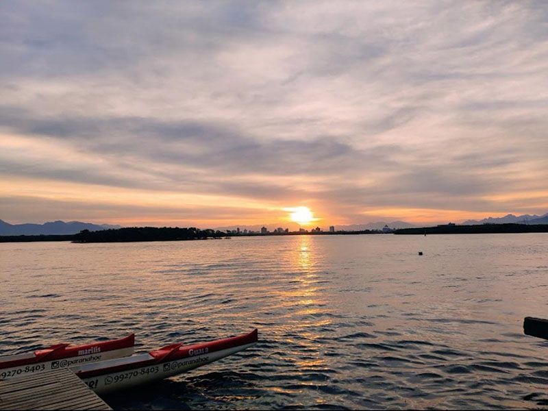 Um pôr do sol sobre águas calmas, com um barco tradicional de balancim em primeiro plano e a costa ao longe.