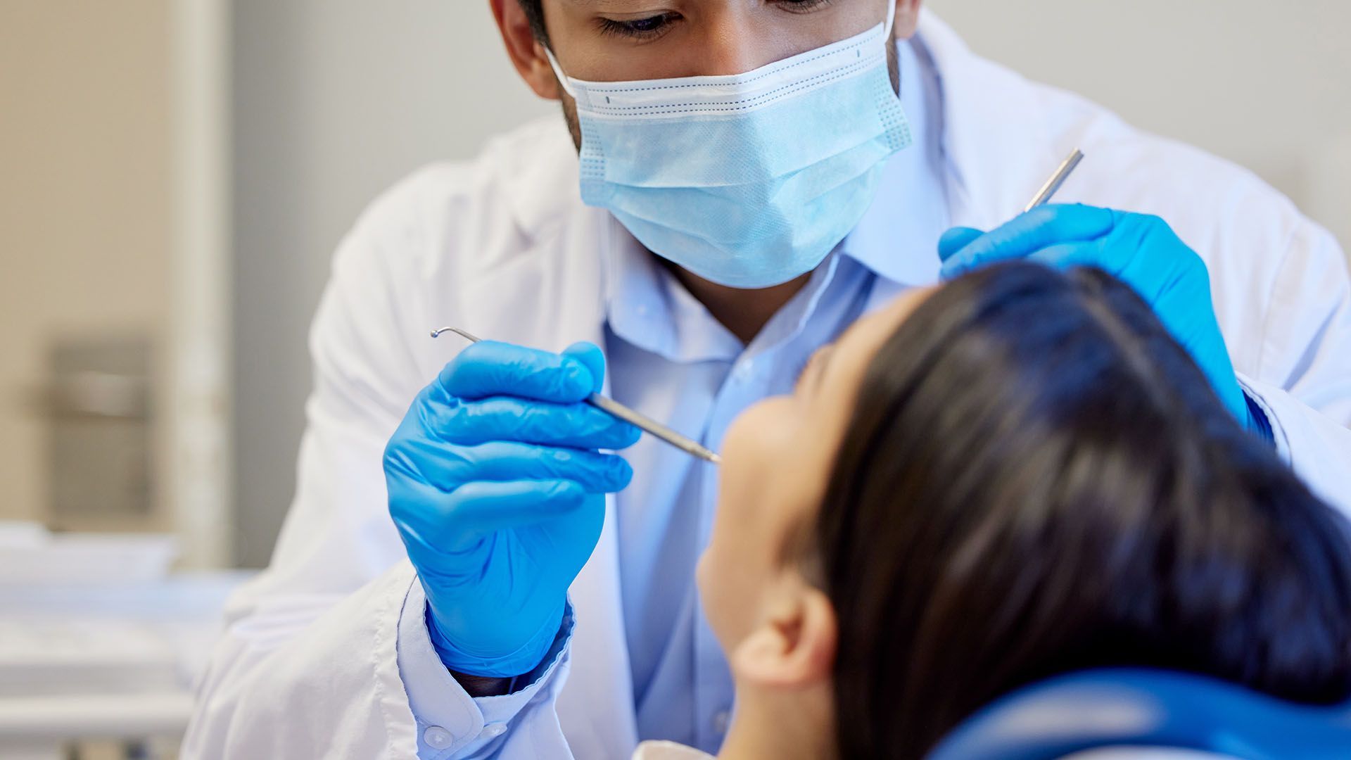 Dentist examining a patient's teeth. The dentist wears a mask, gloves, and a lab coat; the patient is in a dental chair.