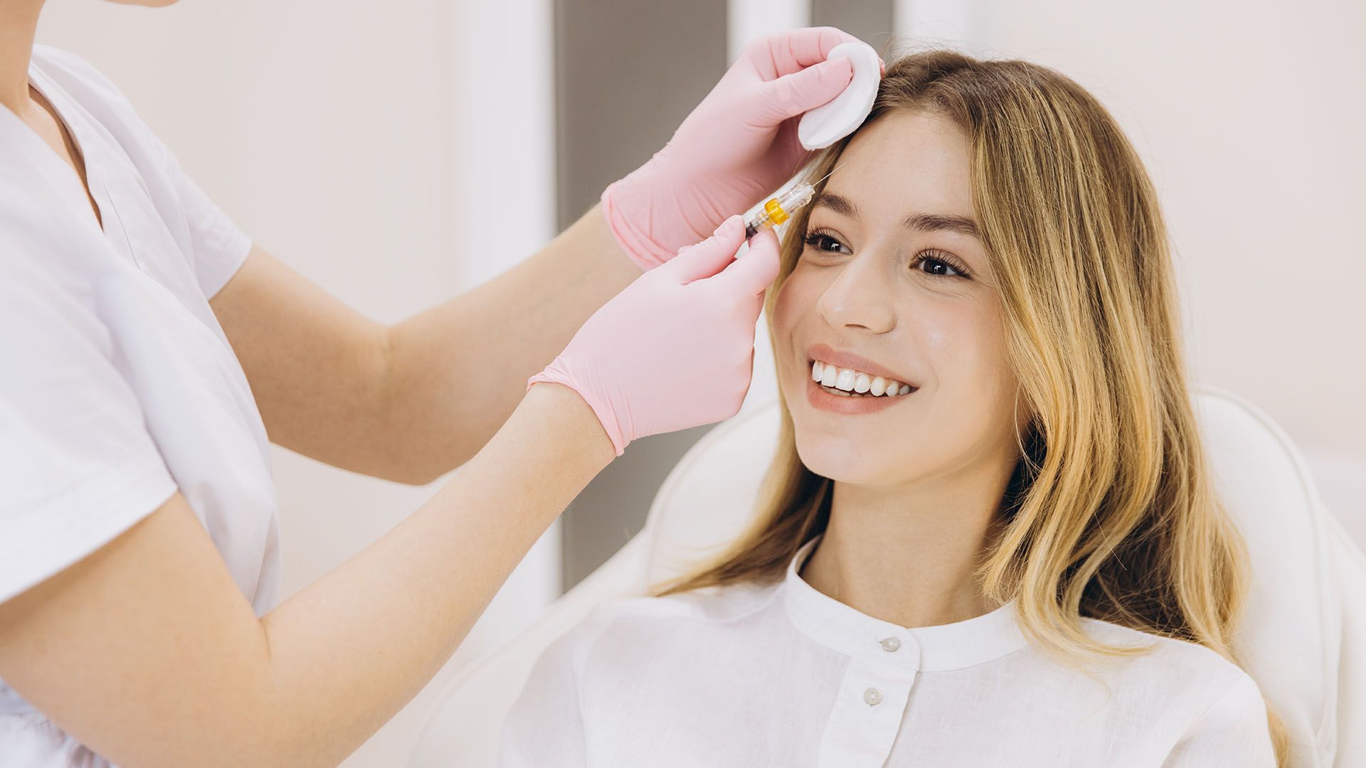 A smiling woman receiving a cosmetic injection on her forehead from a person wearing gloves.
