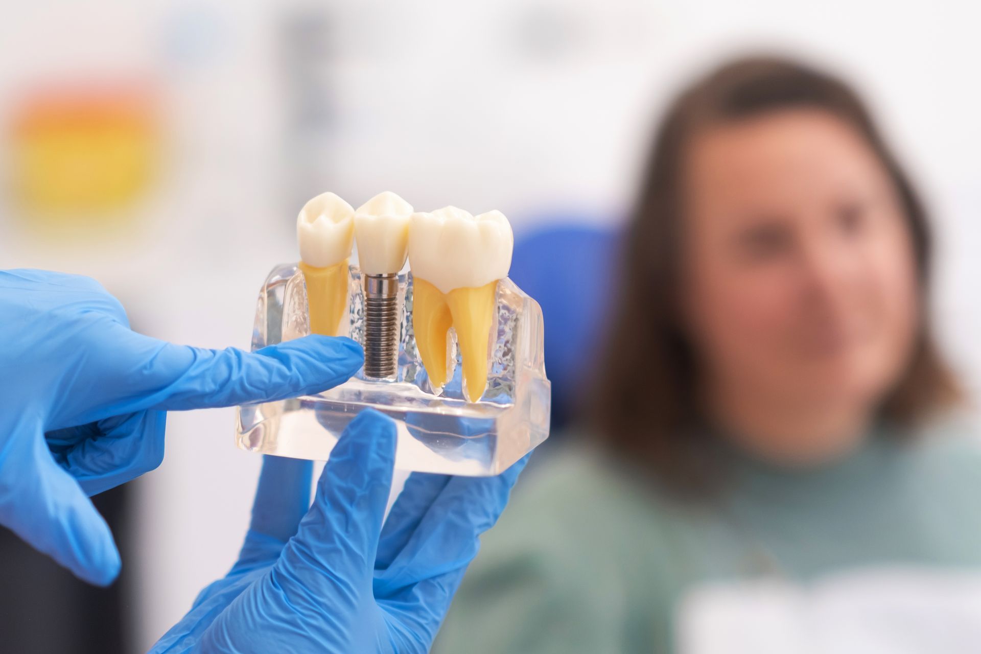A gloved hand holds a dental model demonstrating a tooth implant between two natural teeth, with a patient in the background.