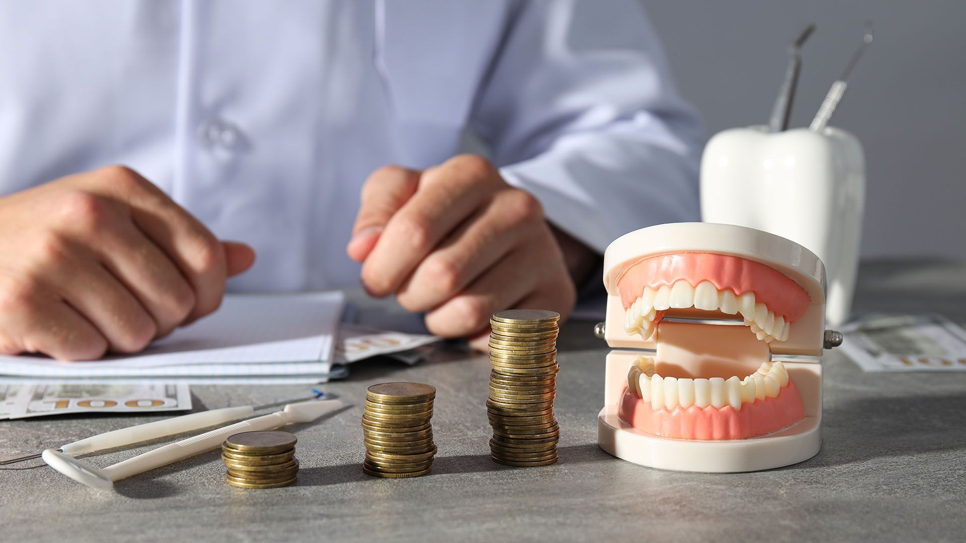 Dentist's hands, dental model, and stacks of coins on a table, symbolizing dental costs.